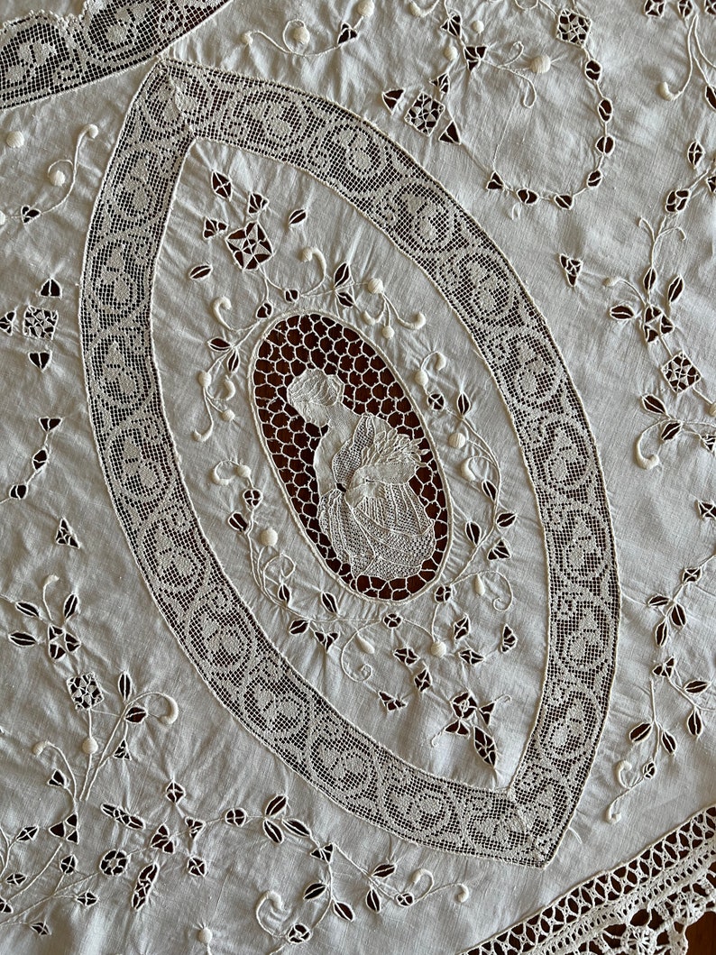 May include: White cotton tablecloth with intricate lace embroidery. The centre features a large oval with a delicate floral design and a smaller oval with a figure in the centre.