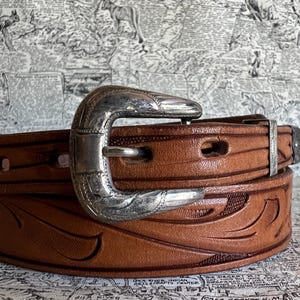 May include: A brown leather belt with a silver-colored buckle. The belt has a carved floral design and is secured with a silver buckle. The belt is a classic western style.