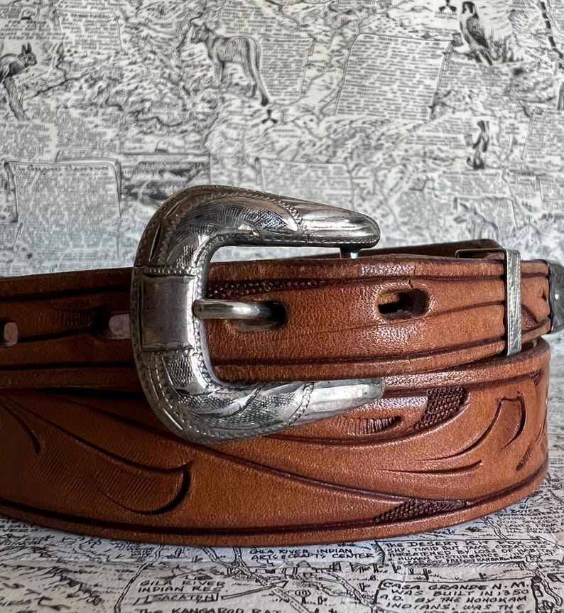 May include: A brown leather belt with a silver-colored buckle. The belt has a carved floral design and is secured with a silver buckle. The belt is a classic western style.
