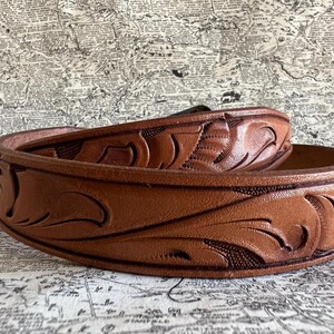 May include: A brown leather belt with a detailed floral pattern. The belt is made of a thick, durable leather and features a classic design. The belt is approximately 1.5 inches wide.