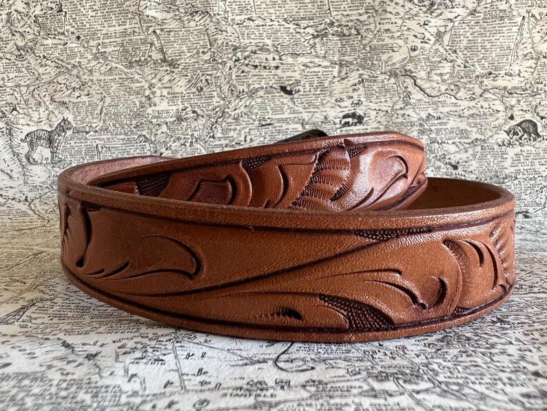 May include: A brown leather belt with a detailed floral pattern. The belt is made of a thick, durable leather and features a classic design. The belt is approximately 1.5 inches wide.