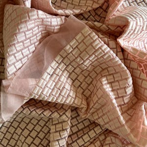 May include: A close-up of a folded, sheer fabric in shades of pink and beige. The fabric has a repeating square pattern, creating a textured effect. The light catches the folds, highlighting the delicate nature of the material. The overall aesthetic is soft and elegant.