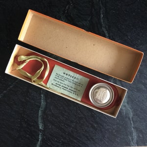 May include: A red rubber ice bag with a yellow rubber strap and a silver cap in a white box. The box has a green label with the text "NOTICE!! Dry threaded sections of the neck and cap of this ICE BAG before storing away to retard oxidation. Keep cap apart from bag when not in use. DAVOL RUBBER COMPANY Providence 2, Rhode Island, U.S.A."