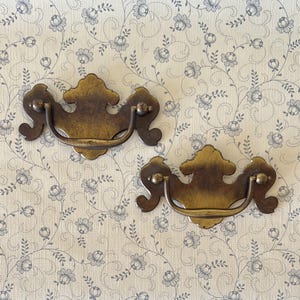 May include: Two matching brass drawer pulls featuring a decorative backplate with a scalloped edge and a curved, swinging bail handle. The metal has a darkened, aged patina finish with visible highlights on the raised edges and smooth surfaces.
