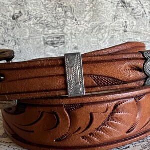 May include: Close-up of a brown leather belt with intricate floral and leaf carvings. The belt features a silver-colored buckle and a decorative silver accent with a leaf design. The leather has a rich, warm tone.