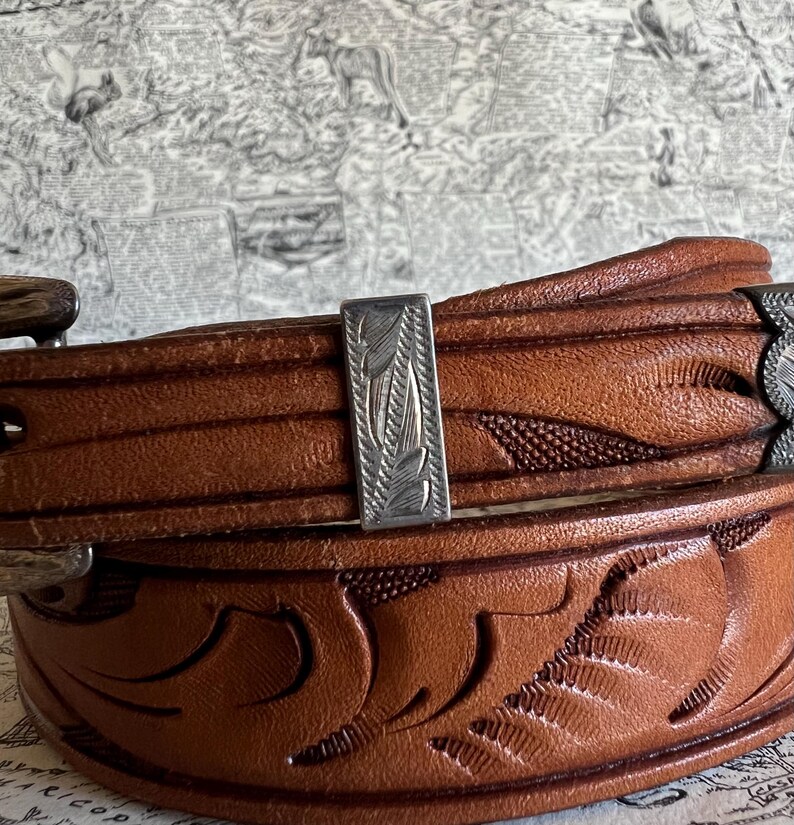 May include: Close-up of a brown leather belt with intricate floral and leaf carvings. The belt features a silver-colored buckle and a decorative silver accent with a leaf design. The leather has a rich, warm tone.