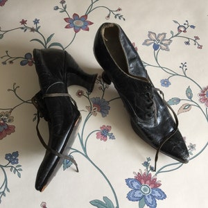 May include: A pair of black leather lace-up shoes with pointed toes and high heels. The shoes are worn and have a vintage look.