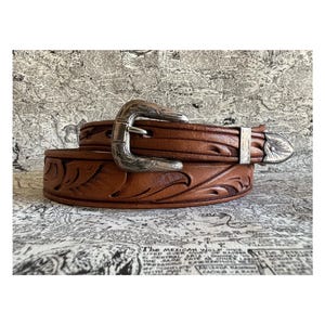 May include: A brown leather belt with a silver buckle and decorative floral tooling. The belt has a classic western style with a detailed design. The buckle and tip are silver-colored, adding a touch of elegance to the accessory.