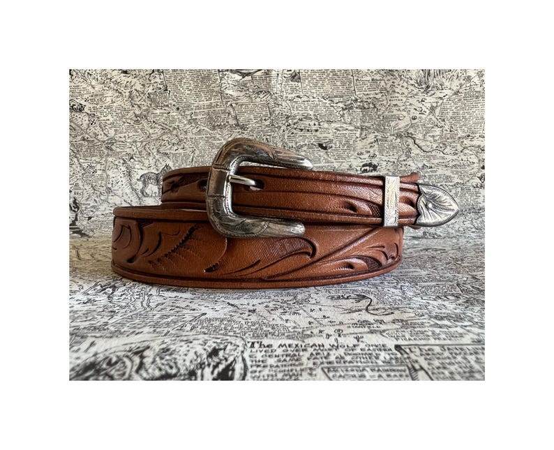 May include: A brown leather belt with a silver buckle and decorative floral tooling. The belt has a classic western style with a detailed design. The buckle and tip are silver-colored, adding a touch of elegance to the accessory.