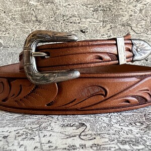 May include: A brown leather belt with a silver buckle and decorative floral tooling. The belt has a classic western style, with a detailed buckle and a matching silver belt loop. The belt is likely made of genuine leather.