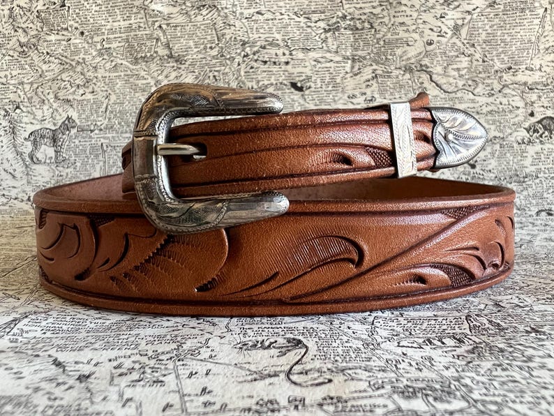 May include: A brown leather belt with a silver buckle and decorative floral tooling. The belt has a classic western style, with a detailed buckle and a matching silver belt loop. The belt is likely made of genuine leather.