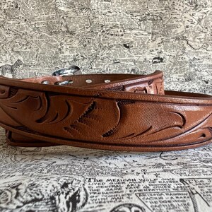 May include: A brown leather belt with intricate carved floral designs. The belt features a silver buckle and multiple holes for adjustment. The leather has a rich, warm tone, and the design is detailed and textured.