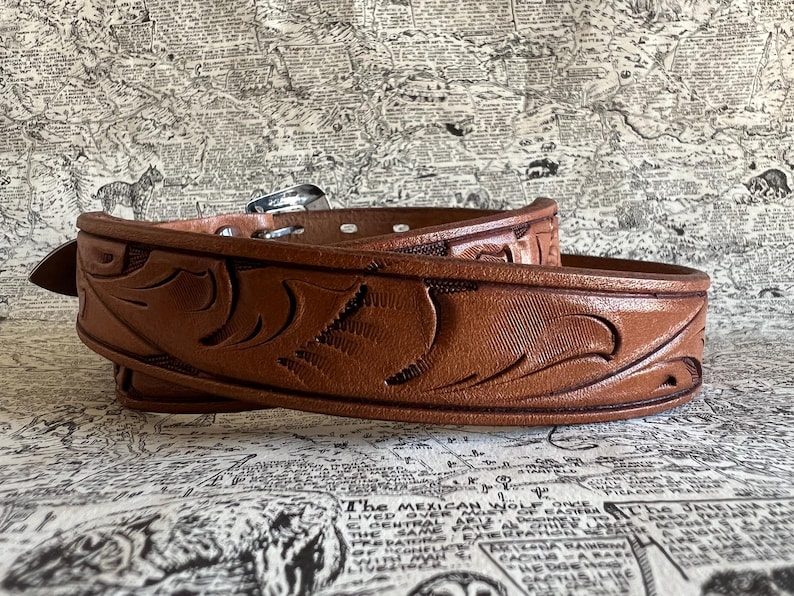 May include: A brown leather belt with intricate carved floral designs. The belt features a silver buckle and multiple holes for adjustment. The leather has a rich, warm tone, and the design is detailed and textured.