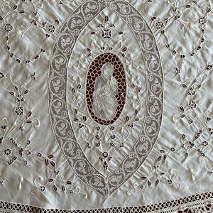 May include: White cotton tablecloth with a delicate lace design featuring a central oval with an embroidered image of a woman. The tablecloth has a scalloped lace border.