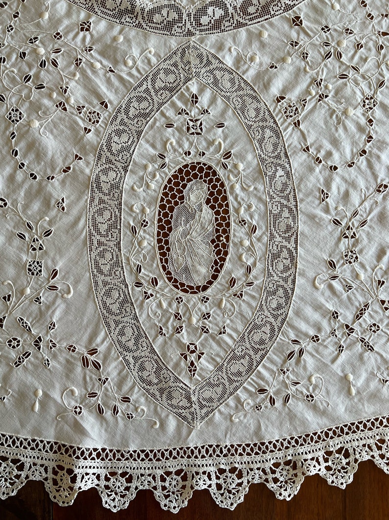 May include: White cotton tablecloth with a delicate lace design featuring a central oval with an embroidered image of a woman. The tablecloth has a scalloped lace border.
