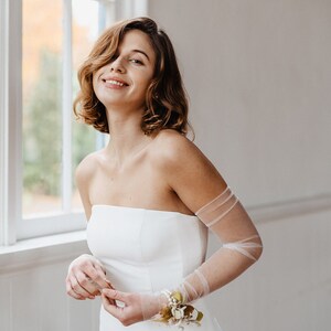May include: A woman wearing a white strapless wedding dress with sheer tulle sleeves and a small floral wrist corsage.