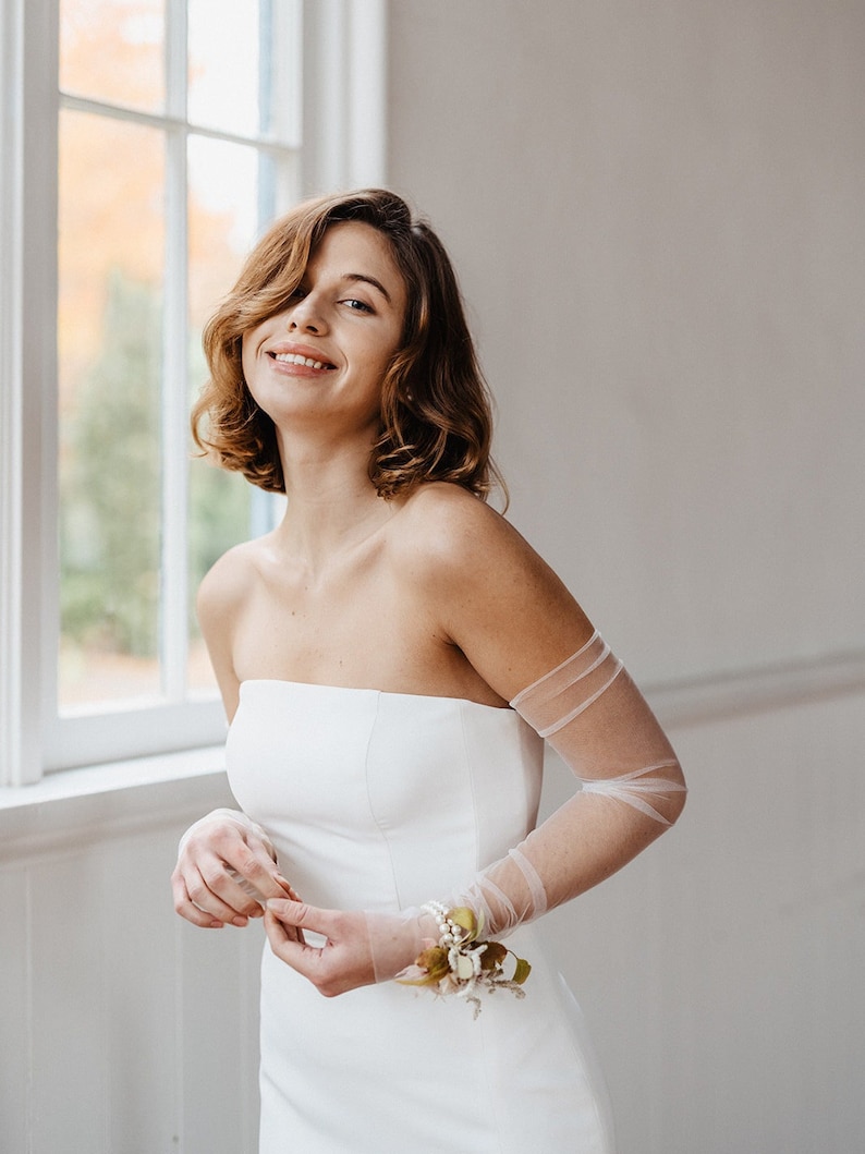 May include: A woman wearing a white strapless wedding dress with sheer tulle sleeves and a small floral wrist corsage.