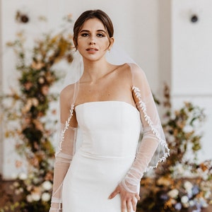 May include: A bride wearing a white strapless wedding dress with a tulle veil and long sleeves. The veil has delicate floral embroidery. The bride is standing in front of a floral backdrop.