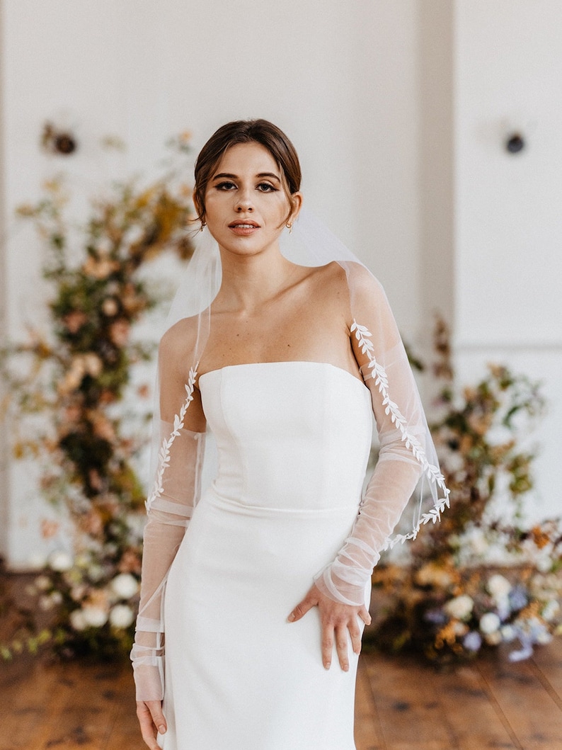 May include: A bride wearing a white strapless wedding dress with a tulle veil and long sleeves. The veil has delicate floral embroidery. The bride is standing in front of a floral backdrop.