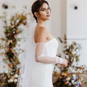 May include: A bride wearing a white strapless wedding dress with sheer long sleeves and a delicate floral veil. The dress has a fitted bodice and a flowing skirt. The bride is standing in a room with a floral backdrop.