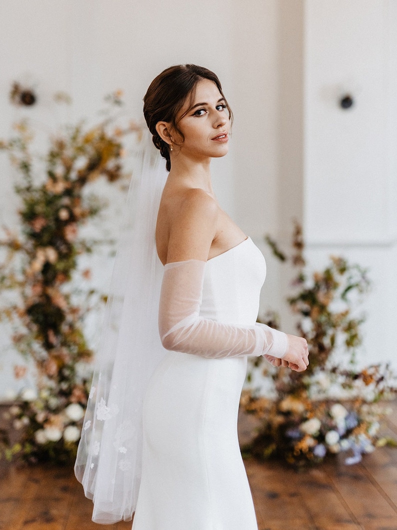 May include: A bride wearing a white strapless wedding dress with sheer long sleeves and a delicate floral veil. The dress has a fitted bodice and a flowing skirt. The bride is standing in a room with a floral backdrop.