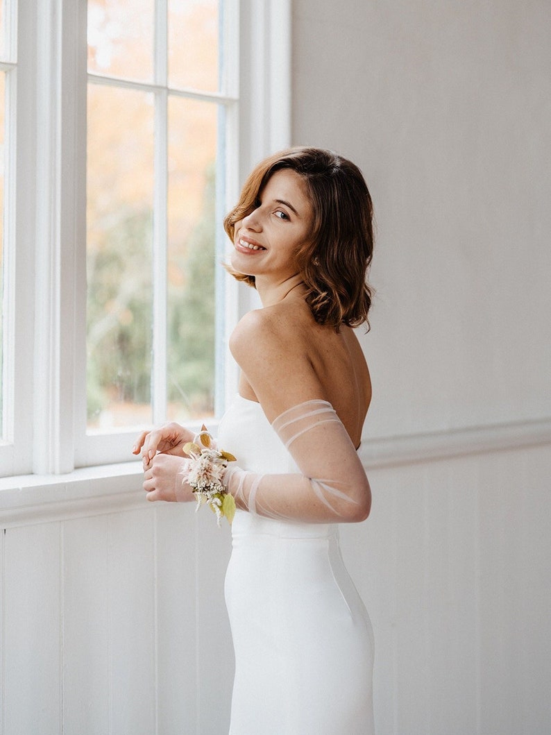 May include: A woman in a white strapless dress with sheer sleeves and a small bouquet of flowers on her wrist. She is standing in front of a window and smiling.
