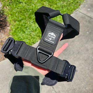 May include: Black tactical dog harness with a black metal D-ring and a black buckle. The harness has a white patch with the text "TACTIPUP" and "Over-Built in the USA".