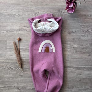 Könnte beinhalten: Ein lila Baby-Overall mit Kapuze und Regenbogen-Applikation. Der Overall hat ein weißes, gemustertes Futter in der Kapuze. Die Regenbogen-Applikation zeigt Farbtöne von Lila, Braun und Weiß. Das Kleidungsstück ist aus einem weichen, strukturierten Material.