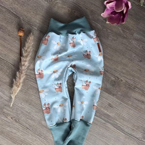Könnte beinhalten: Hellblaue Babyhose mit einem verspielten Aufdruck von Mäusen in Heißluftballons und Gänseblümchen. Die Hose hat einen tealfarbenen Bund und Bündchen. Der Stoff ist weich und das Design ist verspielt.