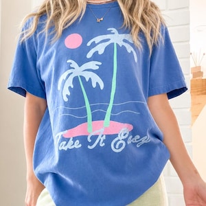 May include: A blue short-sleeve t-shirt featuring a graphic print of two palm trees on a small island, a pink sun, and the text "Take It Easy" in white script. The shirt is paired with light green pants that have a textured appearance.