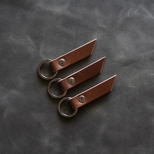 Puede incluir: Tres llaveros de cuero marrón con anillas plateadas y remaches metálicos. Los llaveros tienen forma rectangular con un corte diagonal en el extremo. Están colocados sobre una superficie texturizada de color gris oscuro.