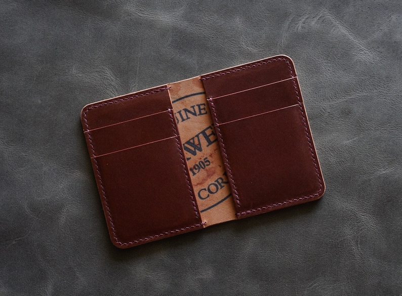 May include: A burgundy leather wallet with multiple card slots and a stitched edge. The inside of the wallet has a brown paper label with the text "Genuine Leather 1905 Corp."