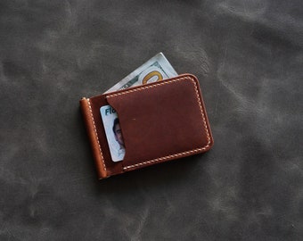 May include: A brown leather money clip wallet with white stitching. A U.S. one hundred dollar bill and a Florida ID card are visible inside the wallet. The wallet is on a dark gray surface.