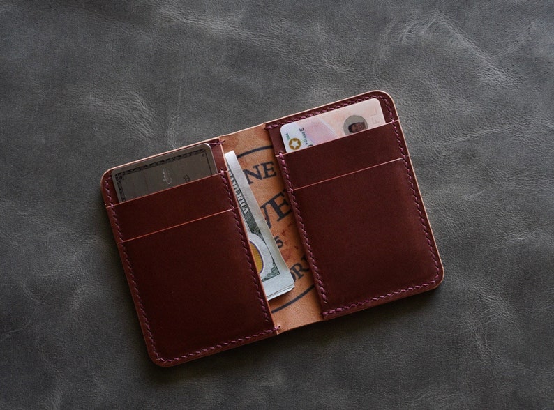 May include: A burgundy leather wallet with multiple card slots and a billfold. The wallet is open and shows a credit card, a driving licence, and a dollar bill.