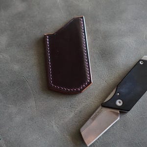 May include: A dark brown leather knife sheath with white stitching next to a black and silver folding knife. The knife blade is open, and the handle has a bottle opener. The knife has the brand name "Kershaw" on the blade.