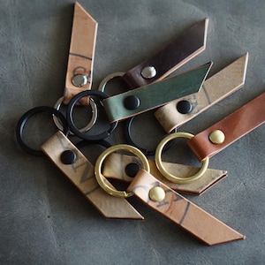 Pode incluir: Uma coleção de chaveiros de couro em várias cores, incluindo marrom, verde e bege. Cada chaveiro apresenta um anel de metal e uma tira de couro com um rebite. Alguns têm anéis pretos, outros dourados.