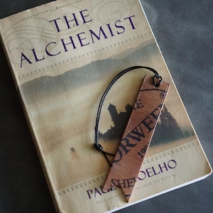 May include: A book titled "The Alchemist" with a brown leather bookmark. The bookmark has the word "ORWELL" printed on it. The book is open, and the bookmark is attached with a black string.