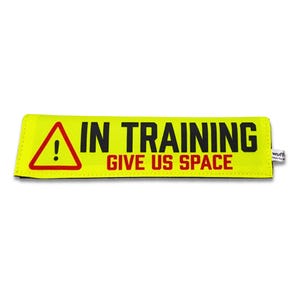 Op de afbeelding: Een rechthoekige neongele stoffen hoes met een rood driehoekig waarschuwingsicoon met een uitroepteken. De tekst IN TRAINING is gedrukt in dikke zwarte letters en de tekst GIVE US SPACE staat eronder in rode letters.