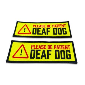 Op de afbeelding: Twee rechthoekige patches met een felgele achtergrond en een zwarte rand. Elke patch bevat een rode driehoek met een uitroepteken, gevolgd door de rode tekst PLEASE BE PATIENT en de zwarte tekst DEAF DOG in vetgedrukte letters.