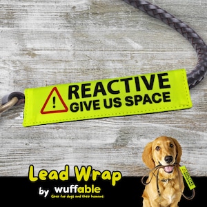 Op de afbeelding: Een neongele hondenriemwikkel met de tekst "REACTIVE GIVE US SPACE" in zwart. De wikkel heeft een rood waarschuwingsdriehoeksymbool. Een bruine en zwarte hondenriem is aan de wikkel bevestigd. De afbeelding toont ook een golden retriever.