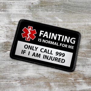 Könnte beinhalten: Schwarz-weißer Patch mit einem roten medizinischen Symbol und dem Text "FAINTING IS NORMAL FOR ME ONLY CALL 999 IF I AM INJURED".