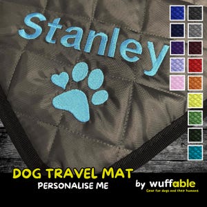 Op de afbeelding: Een grijze, gewatteerde hondenreismat met de naam "Stanley" in lichtblauw geborduurd en een bijpassend pootafdrukontwerp. De mat toont ook de tekst "DOG TRAVEL MAT" en "PERSONALIZE ME".
