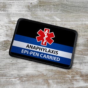 Könnte beinhalten: Schwarzer und blauer Patch mit einem roten medizinischen Symbol und dem Text "Anaphylaxie Epi-Pen getragen".