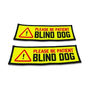 Op de afbeelding: Twee rechthoekige patches met een felgele achtergrond en een zwarte rand. Elke patch bevat een rode driehoek met een uitroepteken, de rode tekst PLEASE BE PATIENT en de zwarte tekst BLIND DOG in een vetgedrukt, schreefloos lettertype.