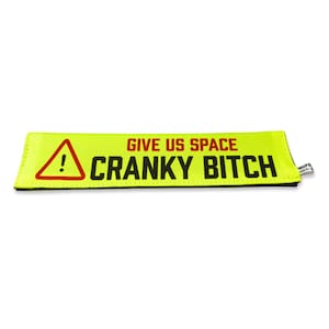 Può includere: Una toppa rettangolare in tessuto giallo neon con un triangolo di avvertimento rosso con un punto esclamativo sul lato sinistro. Il testo "GIVE US SPACE" appare in rosso sopra il testo nero in grassetto "CRANKY BITCH" sulla destra.
