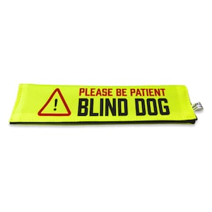 Peut inclure: Une bande de laisse pour chien jaune fluo rectangulaire avec un triangle d'avertissement rouge contenant un point d'exclamation, le texte rouge PLEASE BE PATIENT et le texte noir BLIND DOG. Une petite étiquette blanche est fixée sur le côté.
