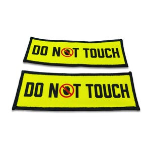 Op de afbeelding: Twee rechthoekige geborduurde patches met een felgele achtergrond en een dikke zwarte rand. De tekst DO NOT TOUCH is gedrukt in dikke zwarte hoofdletters, waarbij een rode cirkel en een handsymbool de letter O vervangen.