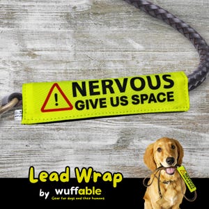 Puede incluir: Una envoltura de correa amarilla con el texto "NERVOUS GIVE US SPACE" y un símbolo de advertencia rojo. La envoltura está unida a una correa trenzada gris. También se ven las palabras "Lead Wrap" y "wuffable".