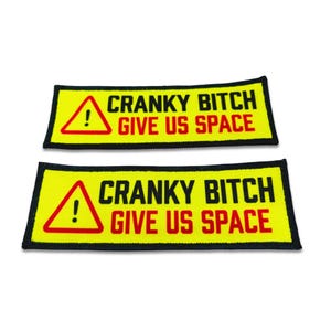 Op de afbeelding: Twee rechthoekige geborduurde patches met een felgele achtergrond en een zwarte rand. Elke patch bevat een rode driehoek met een uitroepteken en de zwarte tekst CRANKY BITCH boven de rode tekst GIVE US SPACE.