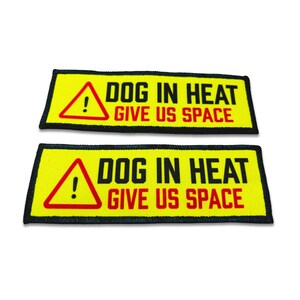 Op de afbeelding: Twee rechthoekige gele geborduurde patches met een zwarte rand. Elke patch heeft links een rode driehoek met een uitroepteken, gevolgd door de zwarte tekst DOG IN HEAT en de rode tekst GIVE US SPACE aan de rechterkant.